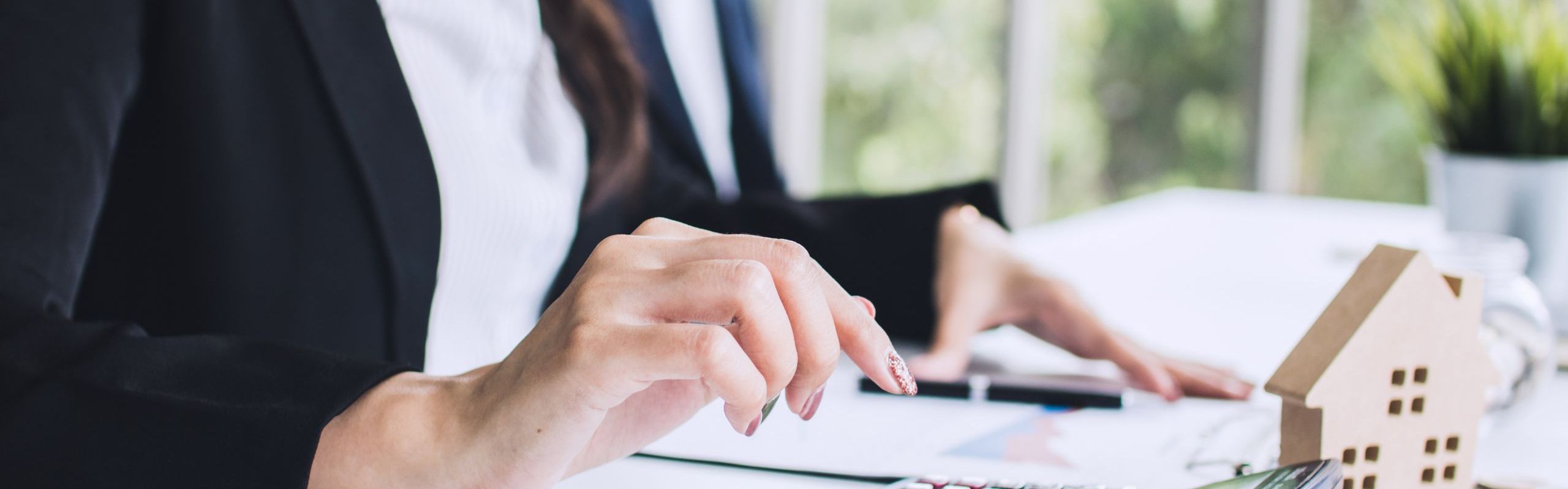 ragazza seduta alla scrivania che digita su una calcolatrice, sulla scrivania ci sono dei fogli, una casetta stilizzata e delle monete