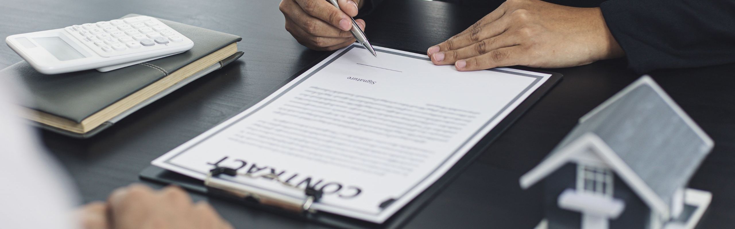Due persone sedute alla scrivania, una scrive su un contratto, scrivania nera, sulla scrivania è posata una casetta in miniatura, un contratto e dei faldoni
