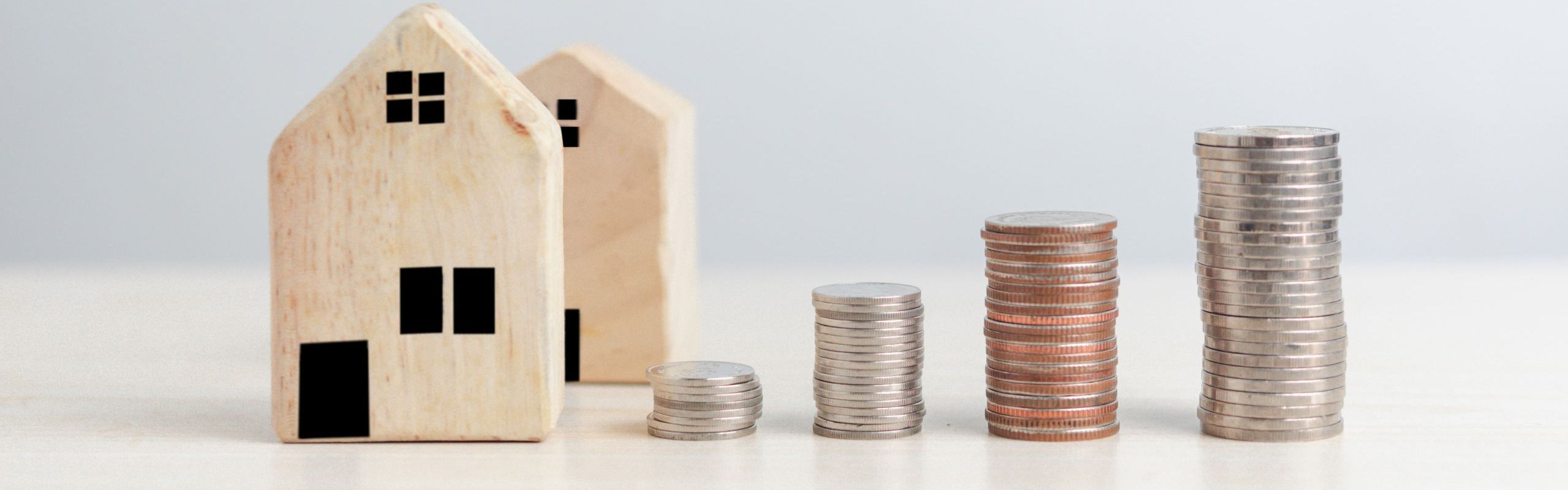 foto di casette stilizzate in legno appoggiate su una scrivania in legno chiaro con a fianco quattro pile di monete