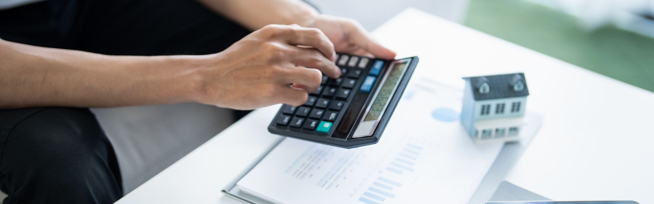 foto di persona con calcolatrice nera in mano su scrivania con fogli stampati con grafici, biro e casetta in miniatura